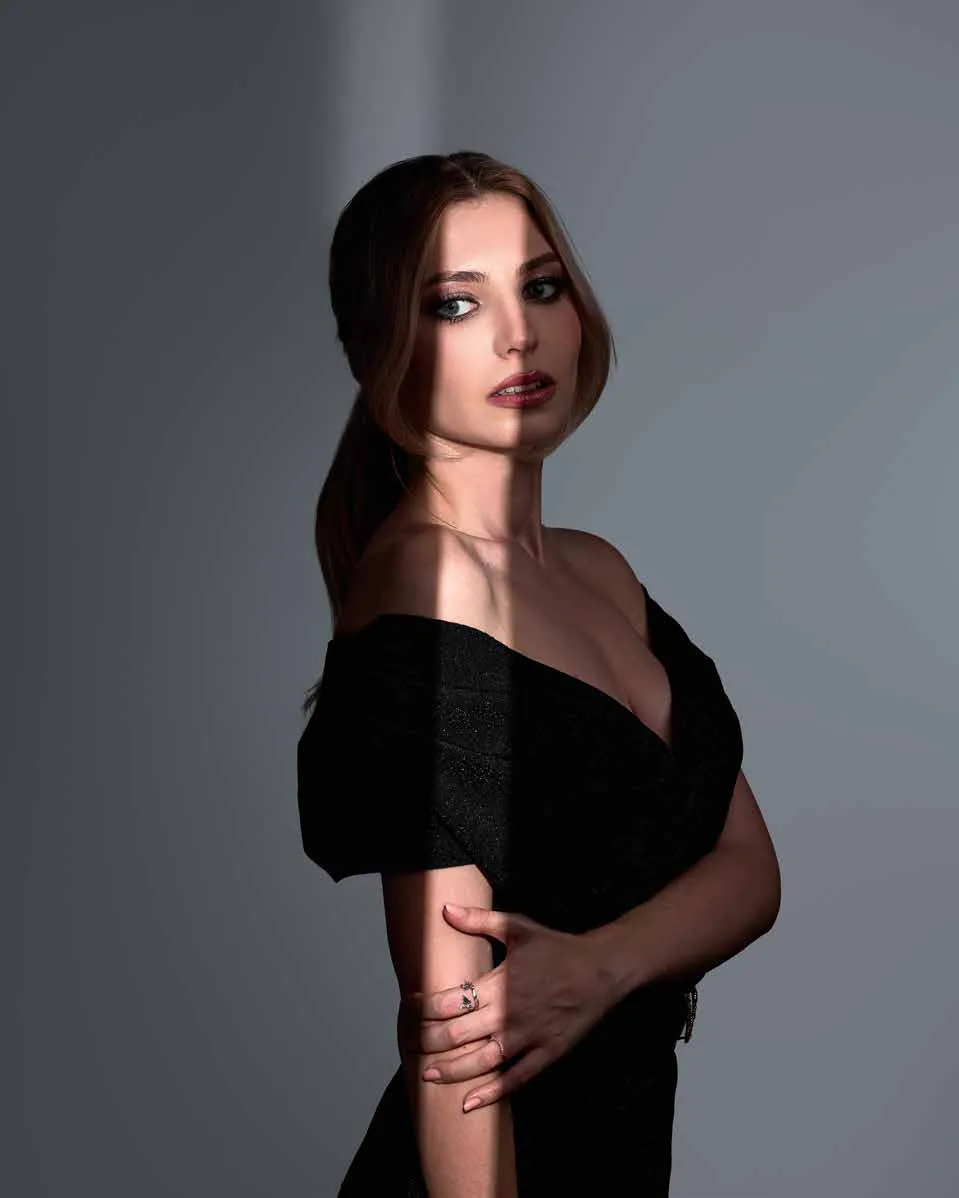 Nicoleta Aliman, portret: rochie neagră cu umerii goi, privire peste umăr, pe un fundal gri, în lumină laterală.