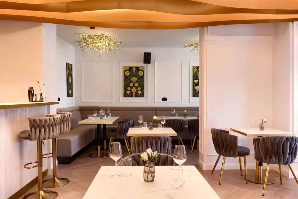 Sală laterală cu banchetă gri-bej, mese albe, tablouri în verde și auriu și un candelabru din ramuri aurii.