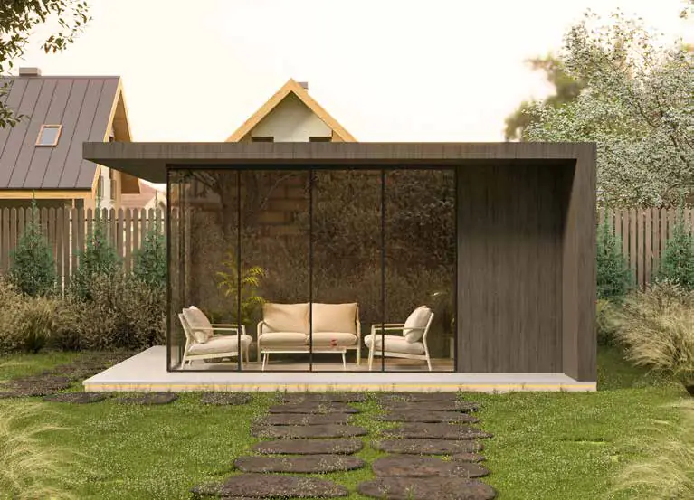 Pavilion de grădină placat cu lemn închis, cu pereți din sticlă și mobilier crem, văzut dinspre poteca de dale.